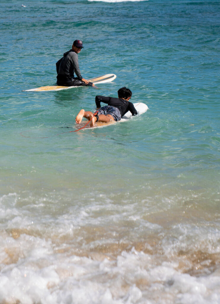 Two people on surfboards in the ocean near the shore, one sitting on a board facing the water, and the other paddling out on their stomach. The water is clear blue with gentle waves.