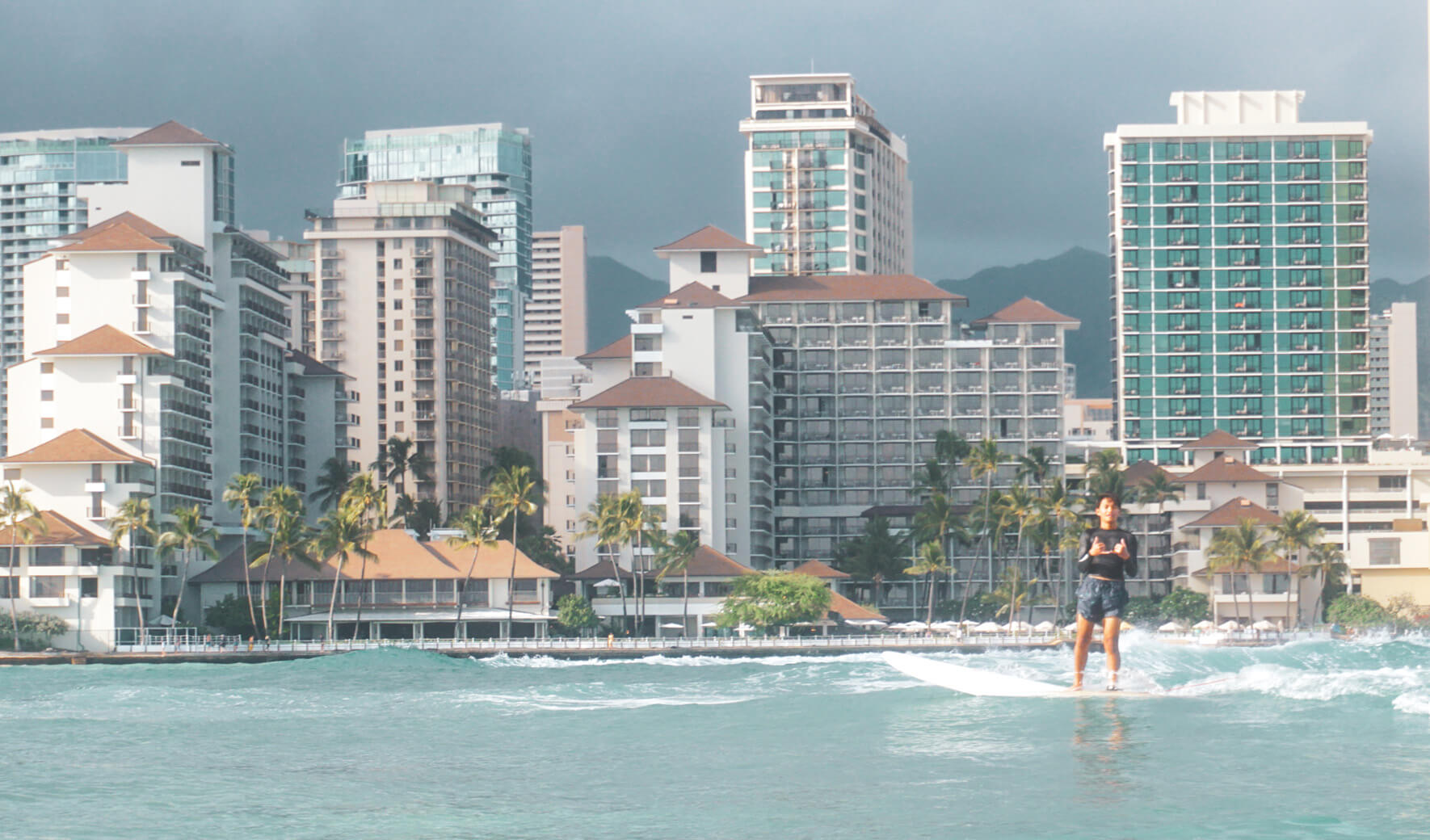 A person surfs on light blue ocean waves with tall hotels and palm trees lining the shore and mountains in the background under a cloudy sky.