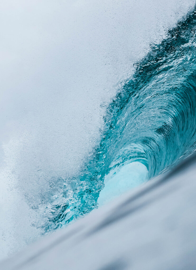 A close-up view of a turquoise ocean wave curling and crashing, with white spray and foam visible against a pale sky.