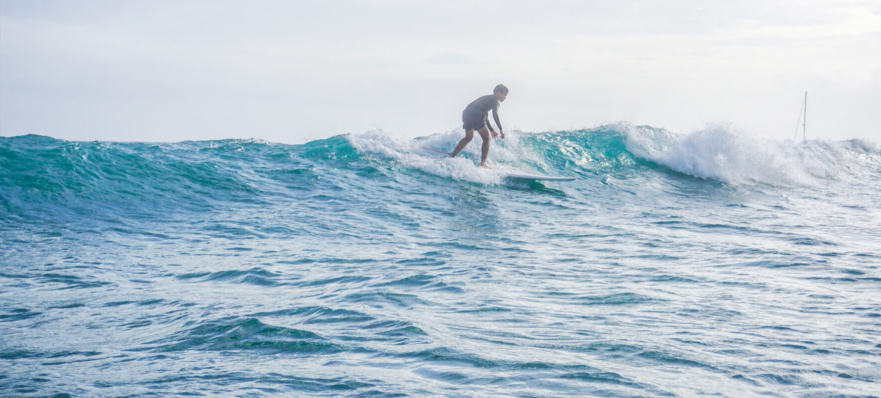 A person surfs on a small wave in the ocean under a bright, slightly cloudy sky. The water is blue and choppy, and the surfer is standing upright on the board.