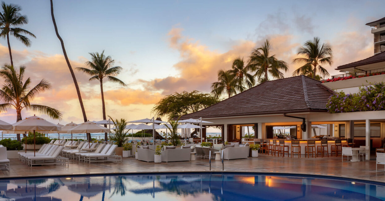 A luxurious resort pool area at sunset, featuring white lounge chairs, umbrellas, a poolside bar with seating, palm trees, and views of the ocean in the background.
