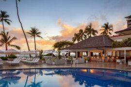 A luxurious resort pool area at sunset, featuring white lounge chairs, umbrellas, a poolside bar with seating, palm trees, and views of the ocean in the background.