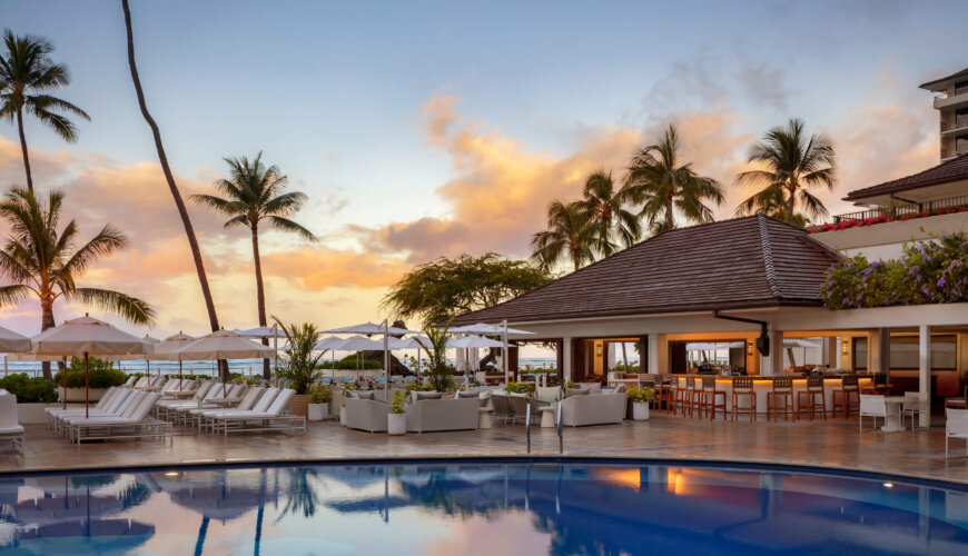 A luxurious resort pool area at sunset, featuring white lounge chairs, umbrellas, a poolside bar with seating, palm trees, and views of the ocean in the background.