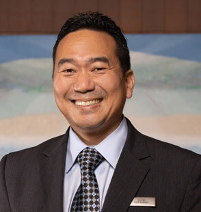 A man in a suit and patterned tie smiles at the camera. He has short dark hair and is wearing a name badge. The background is softly blurred with blue and beige tones.