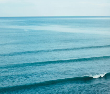 A wide view of the ocean with gentle, evenly spaced waves rolling towards the shore under a pale blue sky. The water is a vibrant blue, and the horizon is visible in the distance.