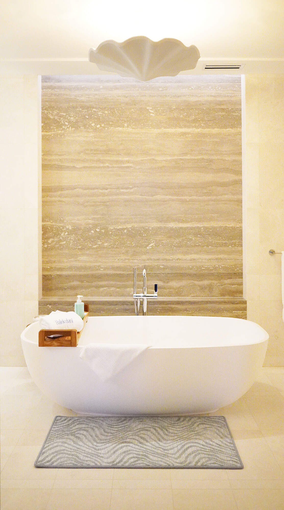 A modern bathroom with a white freestanding bathtub, a wooden bath tray with toiletries, a towel, a striped bath mat, and a beige stone accent wall behind the tub. A shell-shaped ceiling light is overhead.