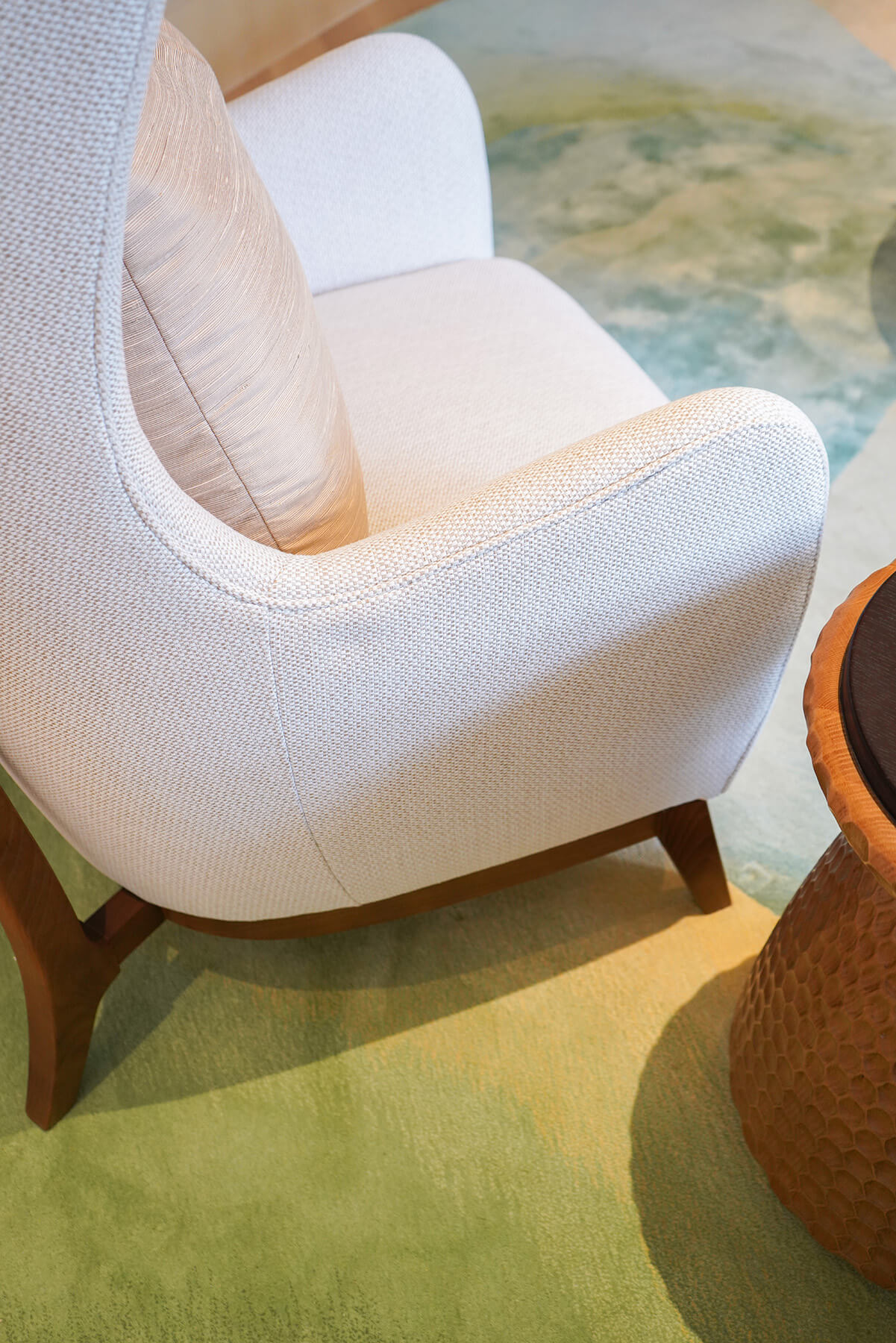 A modern armchair with light upholstery and a beige cushion sits next to a round, textured wooden side table on a green and beige patterned carpet.